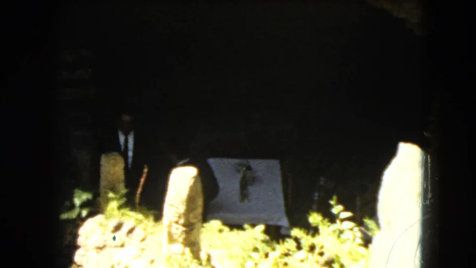 A low, sunlit view shows stone markers, foliage, and a pale covered panel, while the suited figure is mostly hidden by shadow and framing.