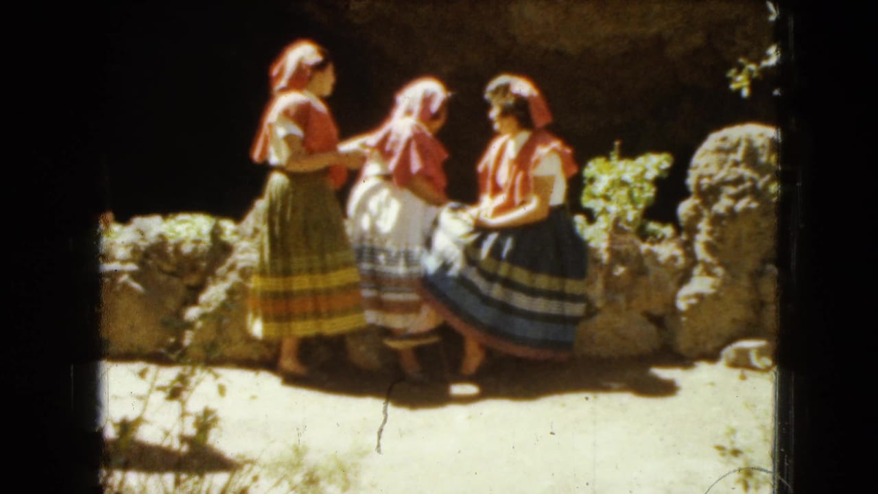 Three women in colorful skirts and headscarves gather beside a low stone wall outdoors.