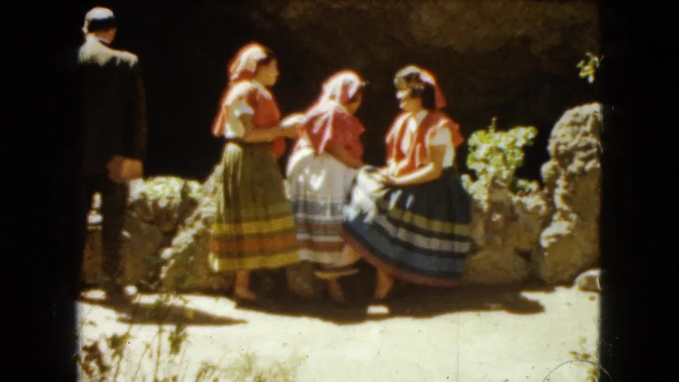 A suited man stands at the edge of the frame while three women in traditional attire gather by a low stone wall in Mexico City footage from 1962.
