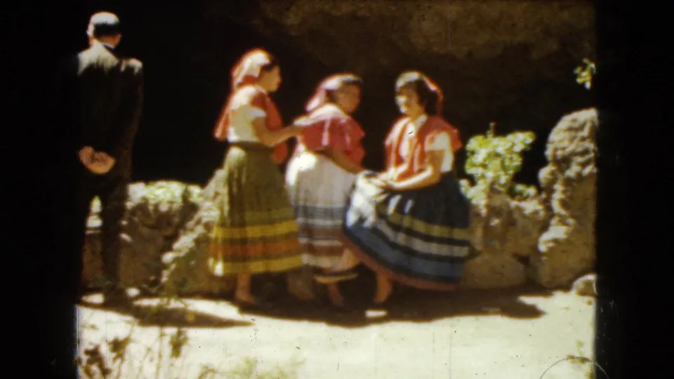 The home-movie frame shows three women in traditional dress near rocks and a low wall, with a man in a dark suit standing to the left.