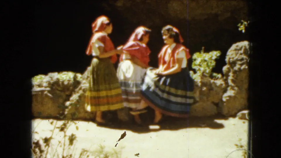 1962 Mexico City 8mm frame showing three young women in traditional dress gathered by a sunlit stone ledge.