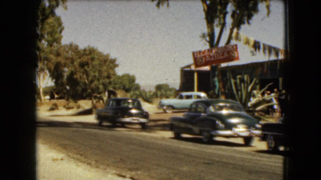 Classic cars pass a roadside restaurant with an El Azteca sign and trees along the street.