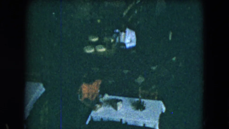 Dark, grainy 8mm frame showing restaurant tables, chairs, and white cloths in a low-light dining area.