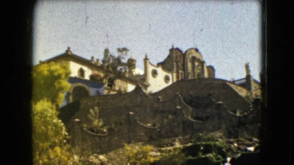 Mexico City travel footage frame with a dark arched foreground structure, pale stucco buildings, trees, and warm 8mm color.