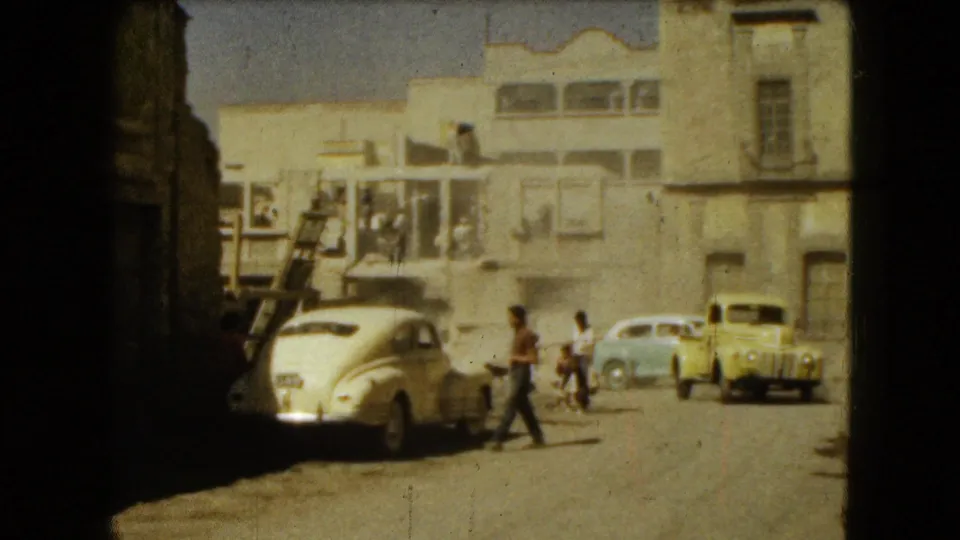 1962 Mexico City street frame with pedestrians, parked mid-century cars, balconies, stucco walls, and a dusty sunlit road.