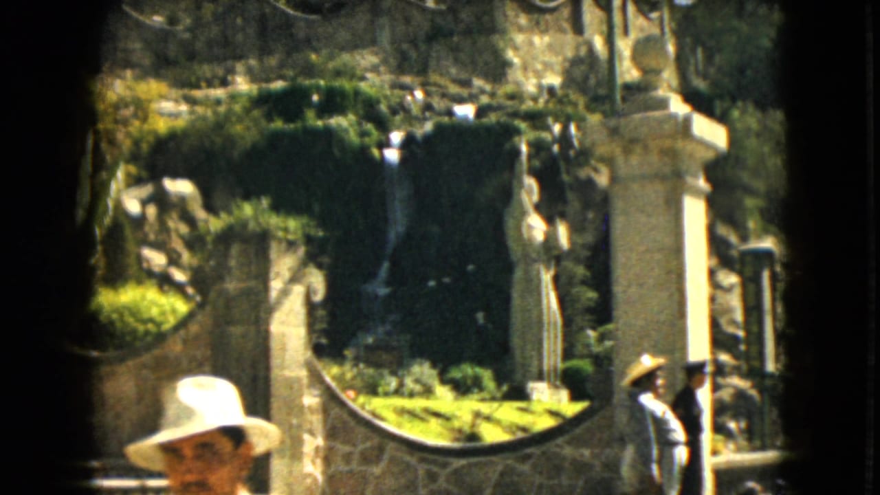 Visitors near stone walls, greenery, a waterfall, and a white statue at an outdoor attraction.