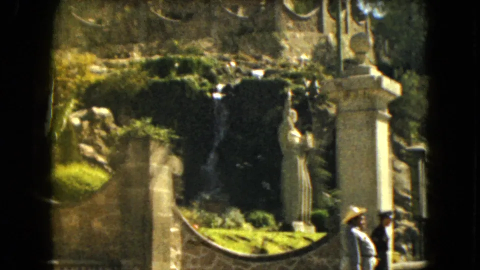 Soft 8mm frame from 1962 Mexico City showing a landscaped waterfall, statue, stonework, and visitors at frame edges.