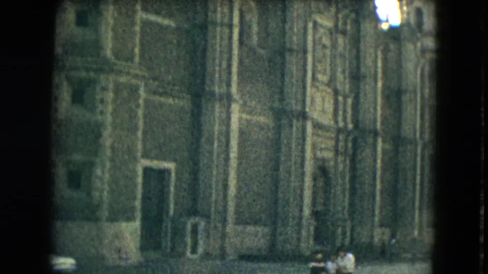 1962 Mexico City home-movie view of an ornate stone building facade, seen from a courtyard with small distant pedestrians near the entrance.