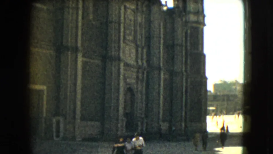 1962 Mexico City home-movie frame showing the side of an ornate building exterior, with distant walkers and a bright opening to the street or plaza.