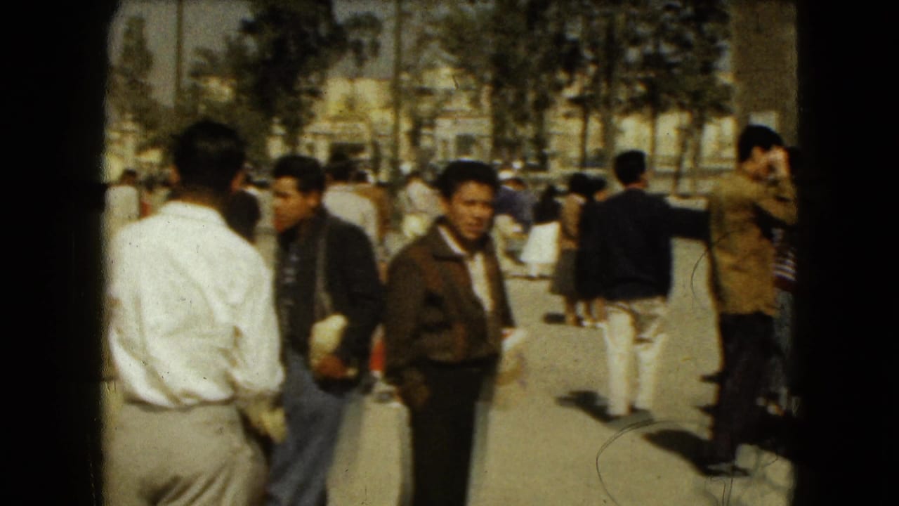 1962 Mexico City home-movie frame showing men in period clothing standing near a street crowd with palm trees and parked cars behind them.