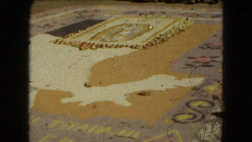 1962 Mexico City home-movie frame focused on an ornate street carpet, possibly devotional, with floral edging and pale shapes on pavement.