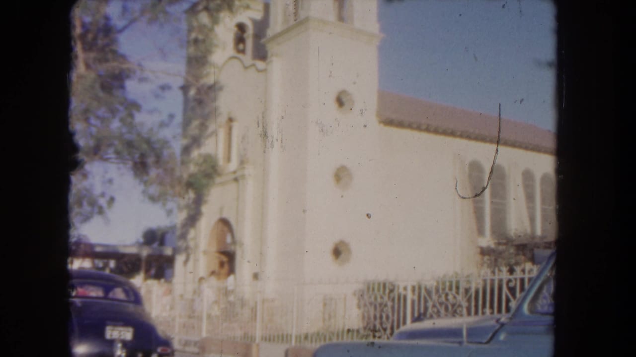 White church with bell tower, arched doorway, fence, trees, and parked cars in bright sun.