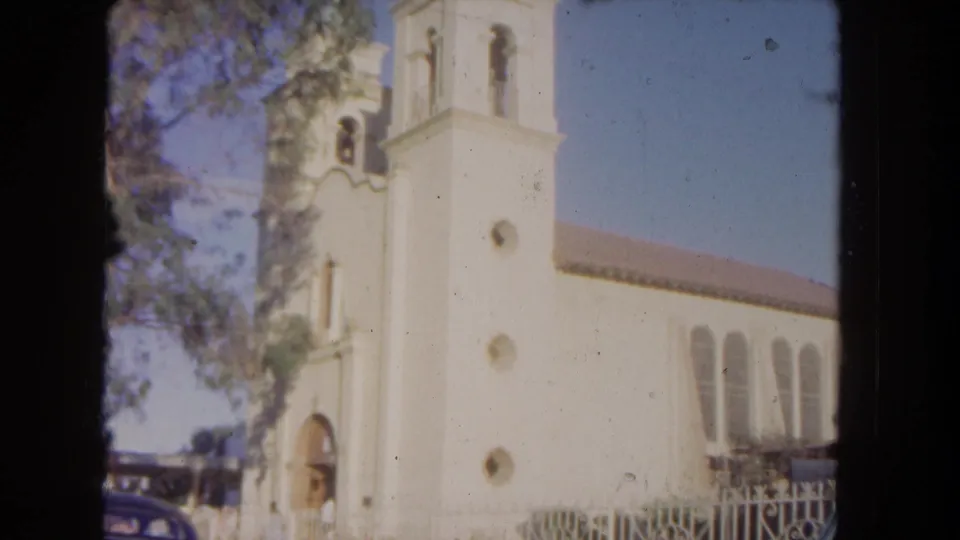 1962 Los Angeles frame of a white church facade with bell tower, arched windows, tree branches, fence, and a partial parked car.