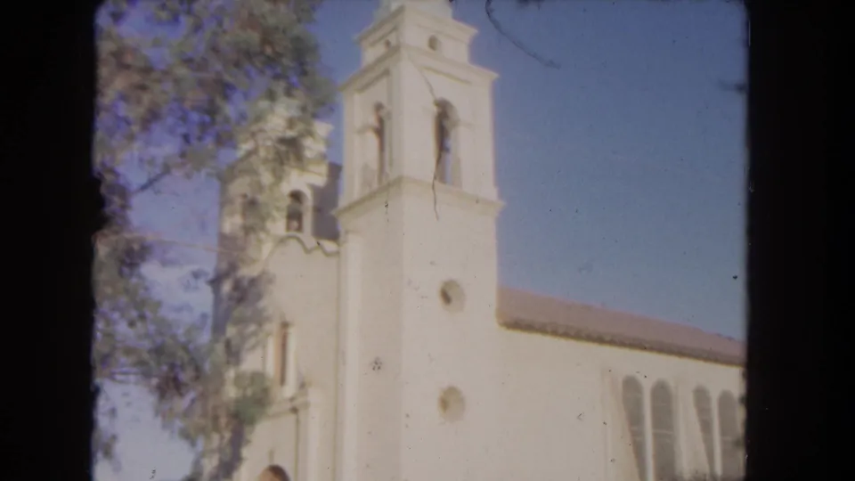 1962 Los Angeles 8mm view of the white church tower and facade, with an arched entrance, side windows, tree branches, fence, and sky.