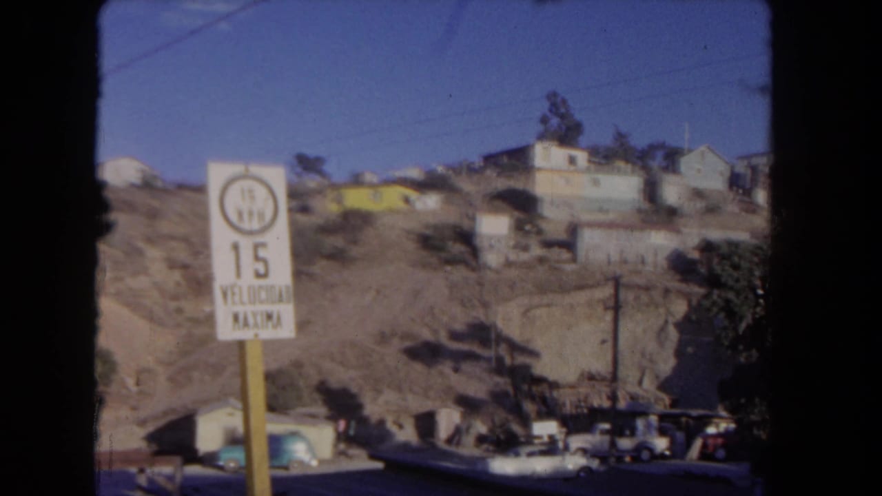 White 15 mph speed limit sign beside a dry hillside with houses and parked cars.