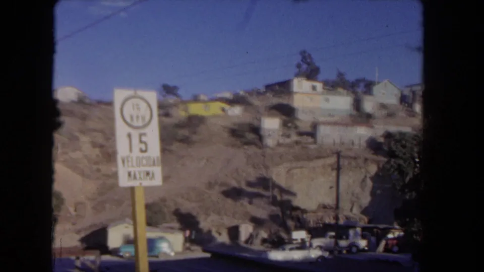 1962 Los Angeles hillside neighborhood with a bilingual 15 mph speed limit sign, dry lots, houses, utility poles, and parked cars.