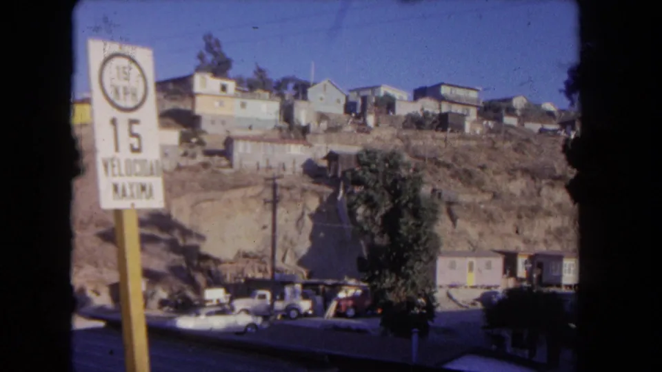 Residential hillside in 1962 Los Angeles, showing a cropped bilingual speed sign, houses on dry ground, parked cars, and utility poles.