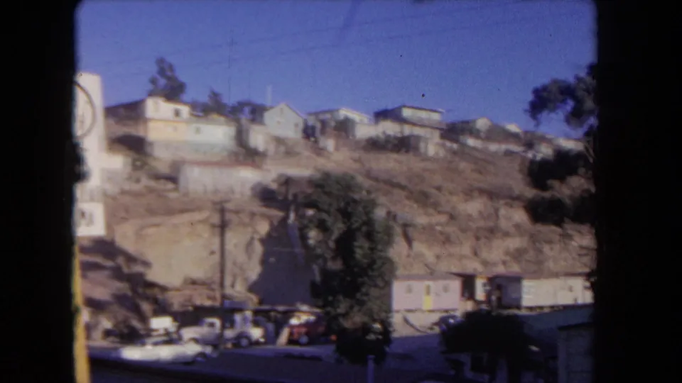 Wide 8mm view of a Los Angeles hillside neighborhood with a partly visible speed sign, dry slope, houses, tree, and parked cars.