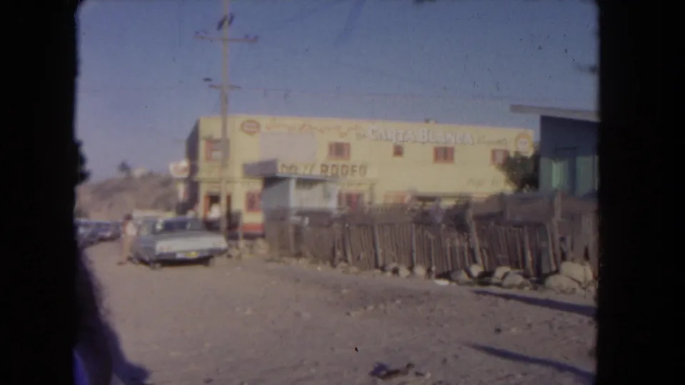 1962 Los Angeles home-movie frame with a yellow signed building, parked sedan, fence, dirt road, and small distant people.
