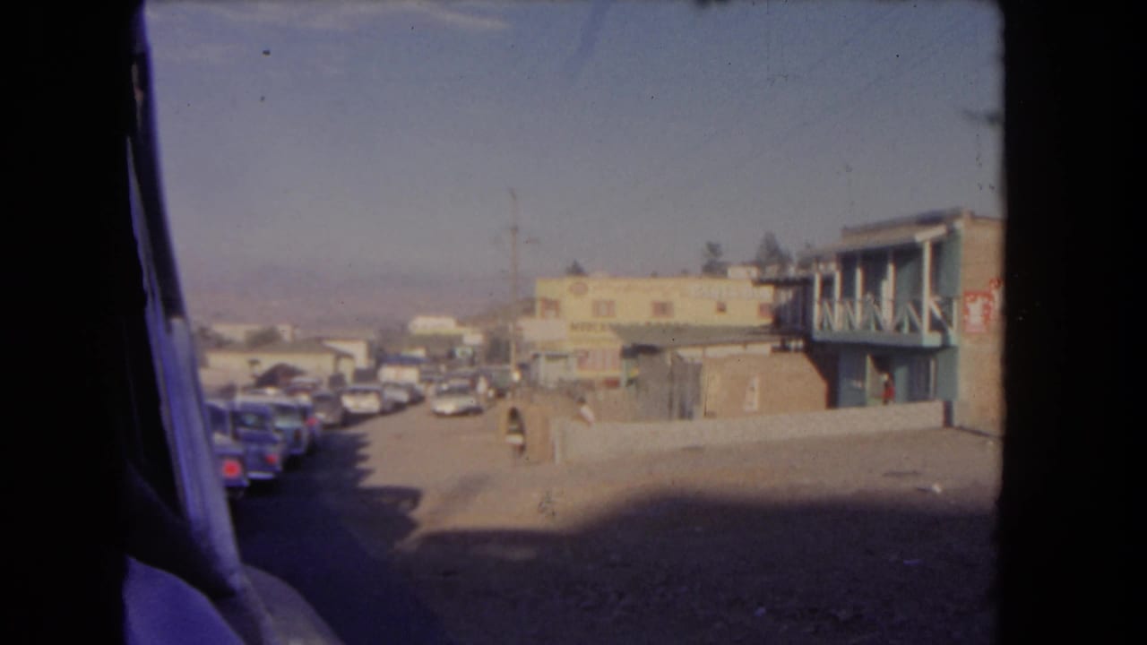 Dusty street viewed from inside a moving car, with parked cars and low-rise buildings ahead.