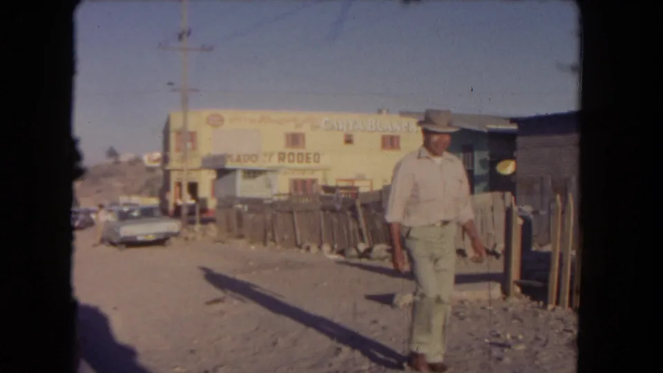 1962 Los Angeles street scene with a man in a hat walking past fencing, parked cars, and readable Carta Blanca and Rodeo signage.