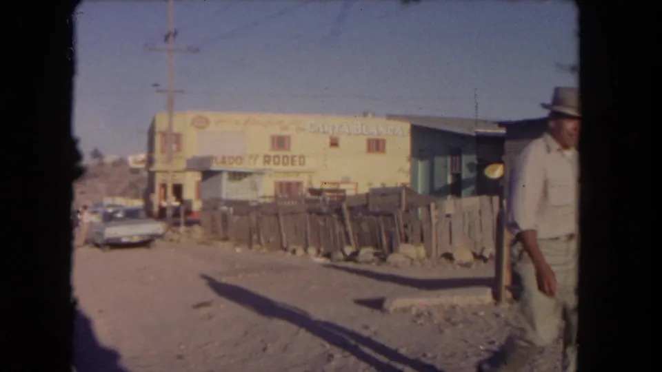 1962 Los Angeles home-movie frame showing a man near the right edge as the car passes a dusty roadside with signs and fencing.