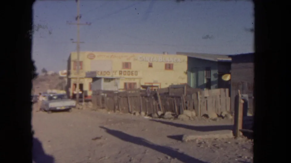 1962 Los Angeles roadside view after the walker passes, showing a dusty street, fence, parked car, utility pole, and commercial signage.