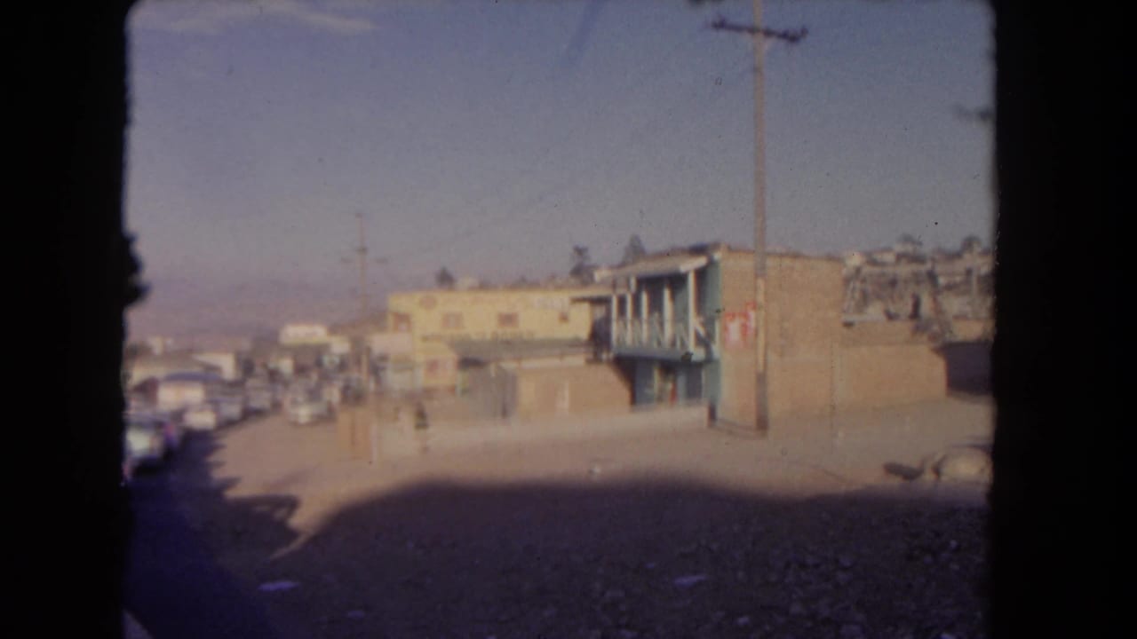 Street seen from a vehicle window with parked cars, low buildings, utility poles, and strong shadow.