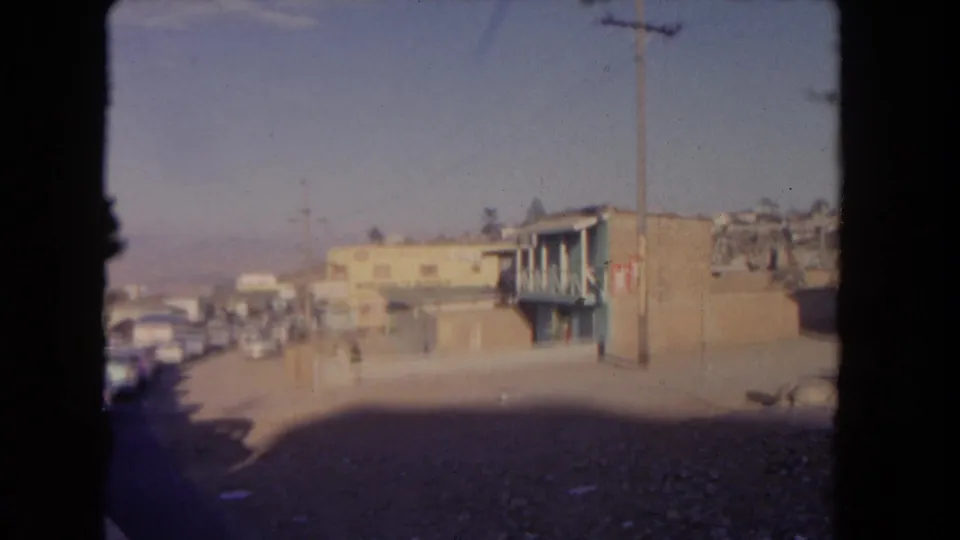 1962 Los Angeles street view from inside a vehicle, showing parked cars, low-rise buildings, utility poles, and bright sun.