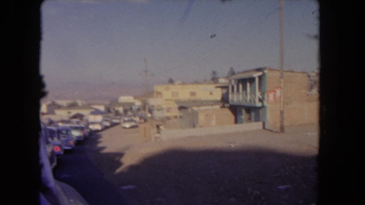 Parked cars line the left side of a street beside low buildings and a balcony structure.