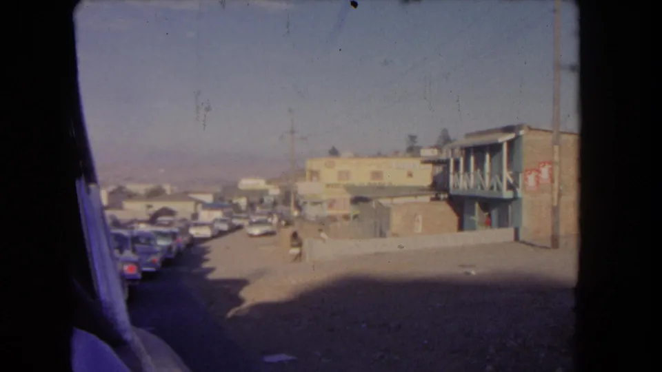 1962 Los Angeles street scene from a moving vehicle, with parked cars, low buildings, balcony railings, and a distant person walking.