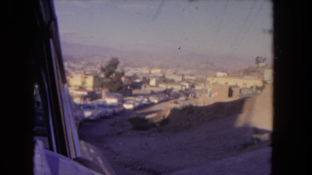 View from a vehicle window over a hilly street with parked cars, buildings, trees, and distant hills.
