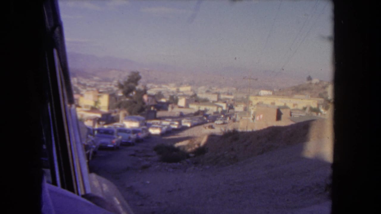 Passenger-side view of a sloping road with parked cars, low buildings, a tree, and hazy hills beyond.