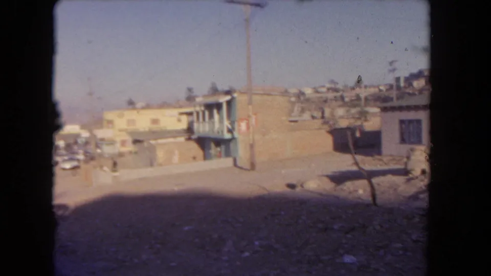 1962 Los Angeles frame showing a low-rise roadside building, parked cars, hillside structures, a utility pole, and heavy foreground shadow.