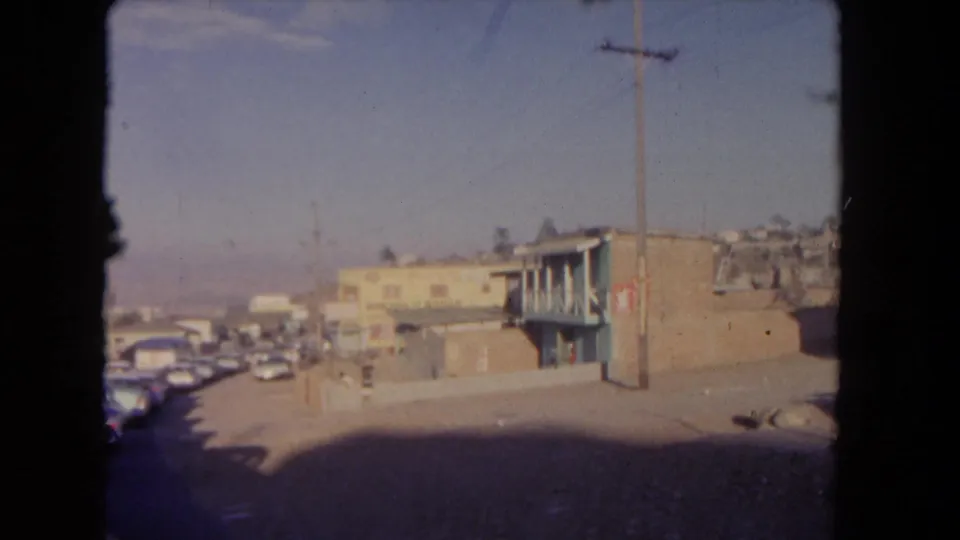 1962 Los Angeles home-movie view of a street corner with a low building, utility pole, parked cars, hillside homes, and clear sky.