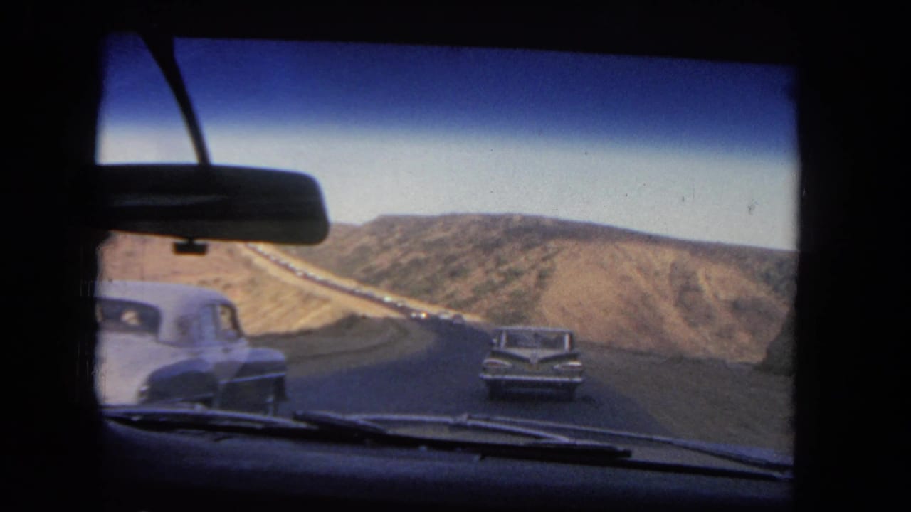 Windshield view in Los Angeles, 1962, with an oncoming white car passing and another sedan ahead on a dry hillside highway.