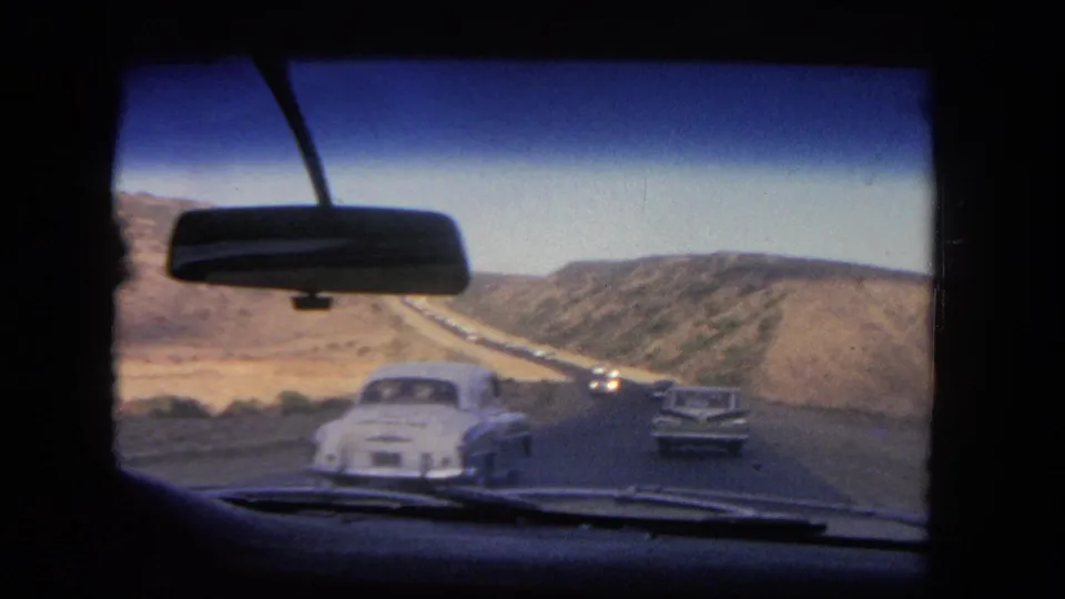 1962 Los Angeles windshield view as a white car passes in the foreground and a sedan continues along the hillside highway.