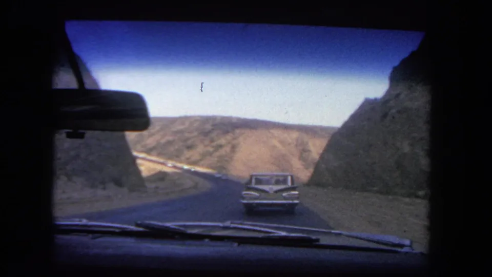 1962 in-car view of a vintage sedan ahead on a canyon road near Los Angeles, with dry hills and dark rock faces around the roadway.