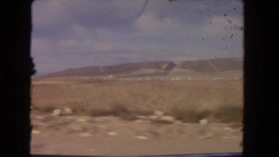 1962 Los Angeles-area 8mm frame showing dry roadside terrain, low mesa ridges, and pale blue sky from a moving car.