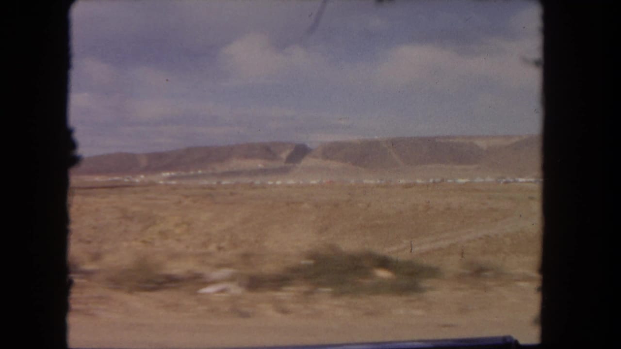 Moving 8mm view of a dry Southern California roadside with low mesa slopes and scattered scrub under a blue sky.