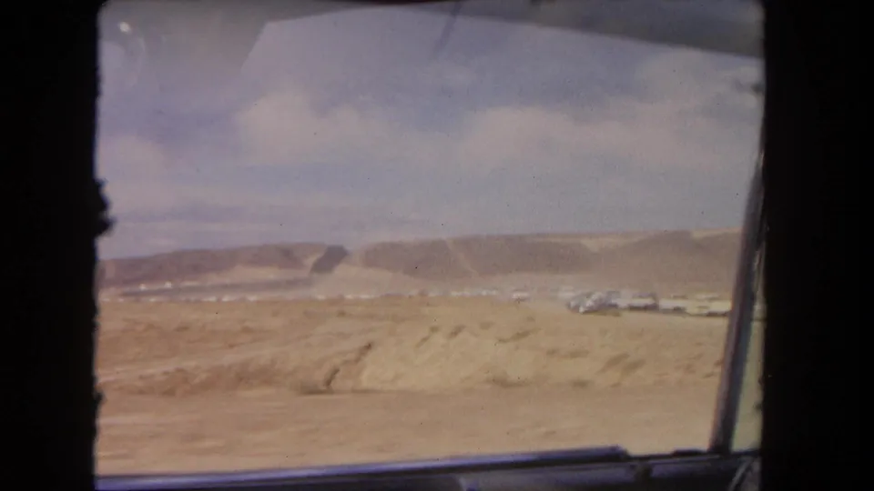 1962 Los Angeles-area roadside view through a vehicle window, with dry ground, mesa slopes, and distant parked cars.