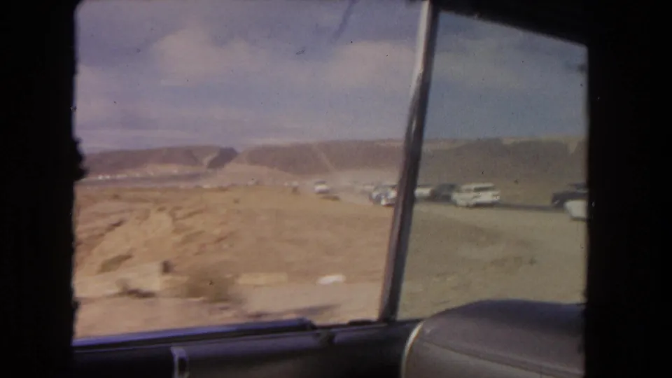 Final 8mm frame from inside a moving vehicle, looking past the window frame toward parked cars, dry ground, and mesa ridges.