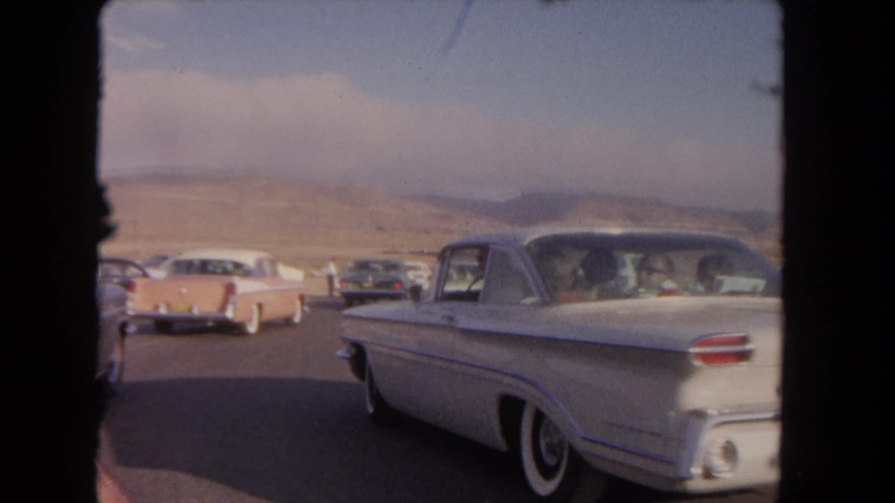 1962 8mm frame of a white classic car on a desert road near Los Angeles, with other period vehicles and mountains beyond.