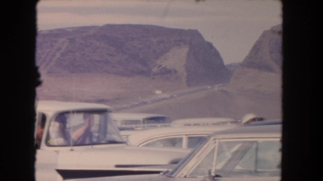 White mid-century car and other vehicles on a desert road with rocky hills behind them.