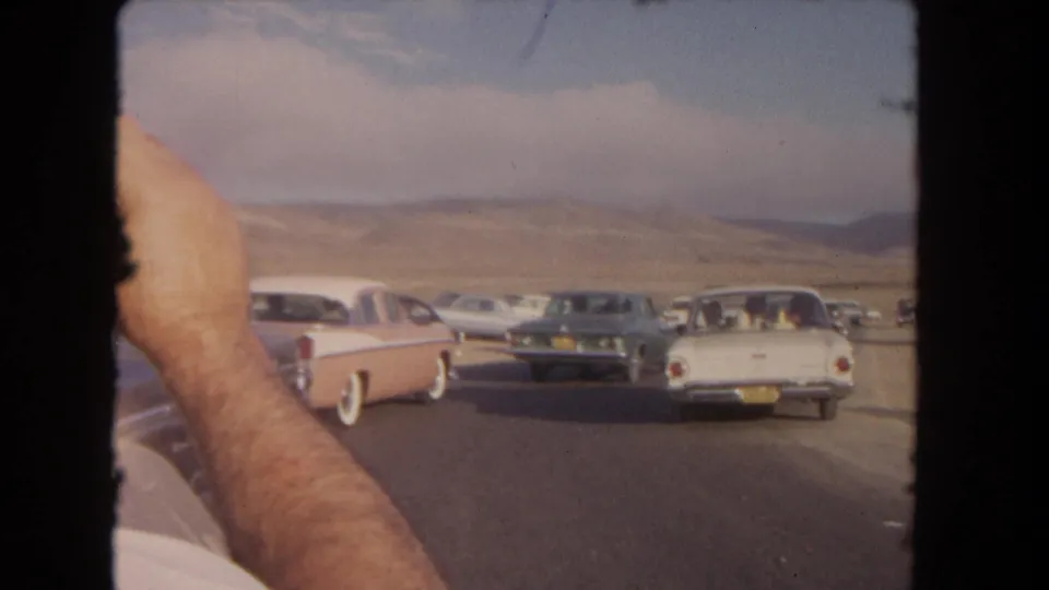 Mid-century cars continue down a desert road in a 1962 8mm California home movie, with a passenger arm visible in the foreground.
