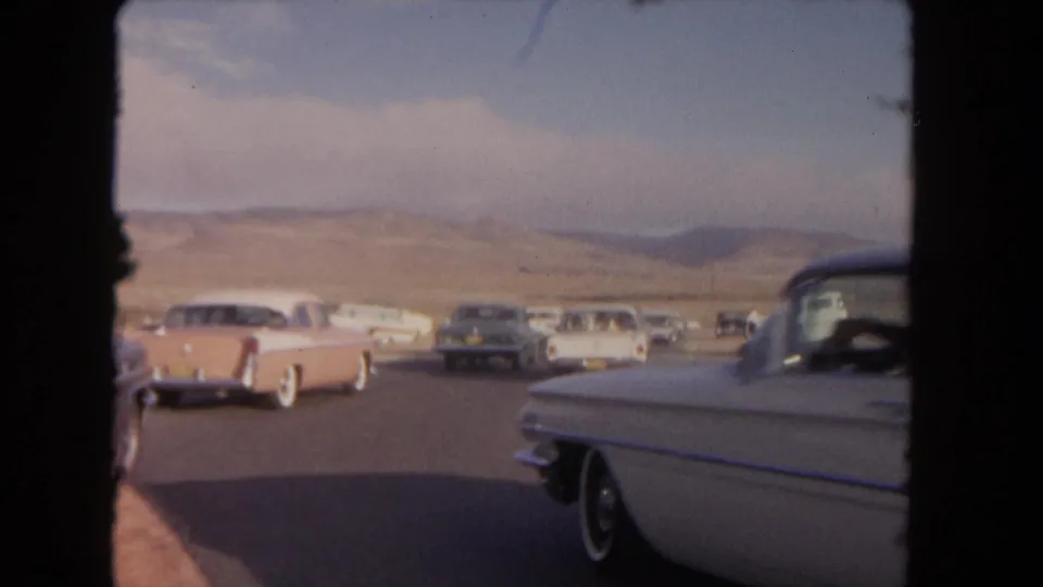 A moving-vehicle view shows several mid-century cars on a desert road in 1962 California footage, with hills and blue sky in the distance.