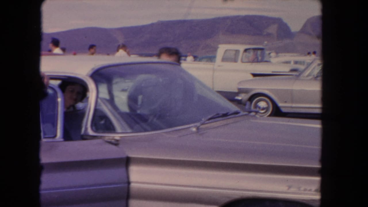 Classic cars in a parking lot with a man partly visible behind a foreground car and mountains beyond.