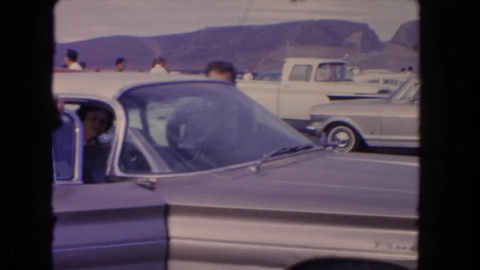 1962 Los Angeles-area 8mm frame showing classic cars in a parking lot, a man near the foreground windshield, people in back, and mountains beyond.