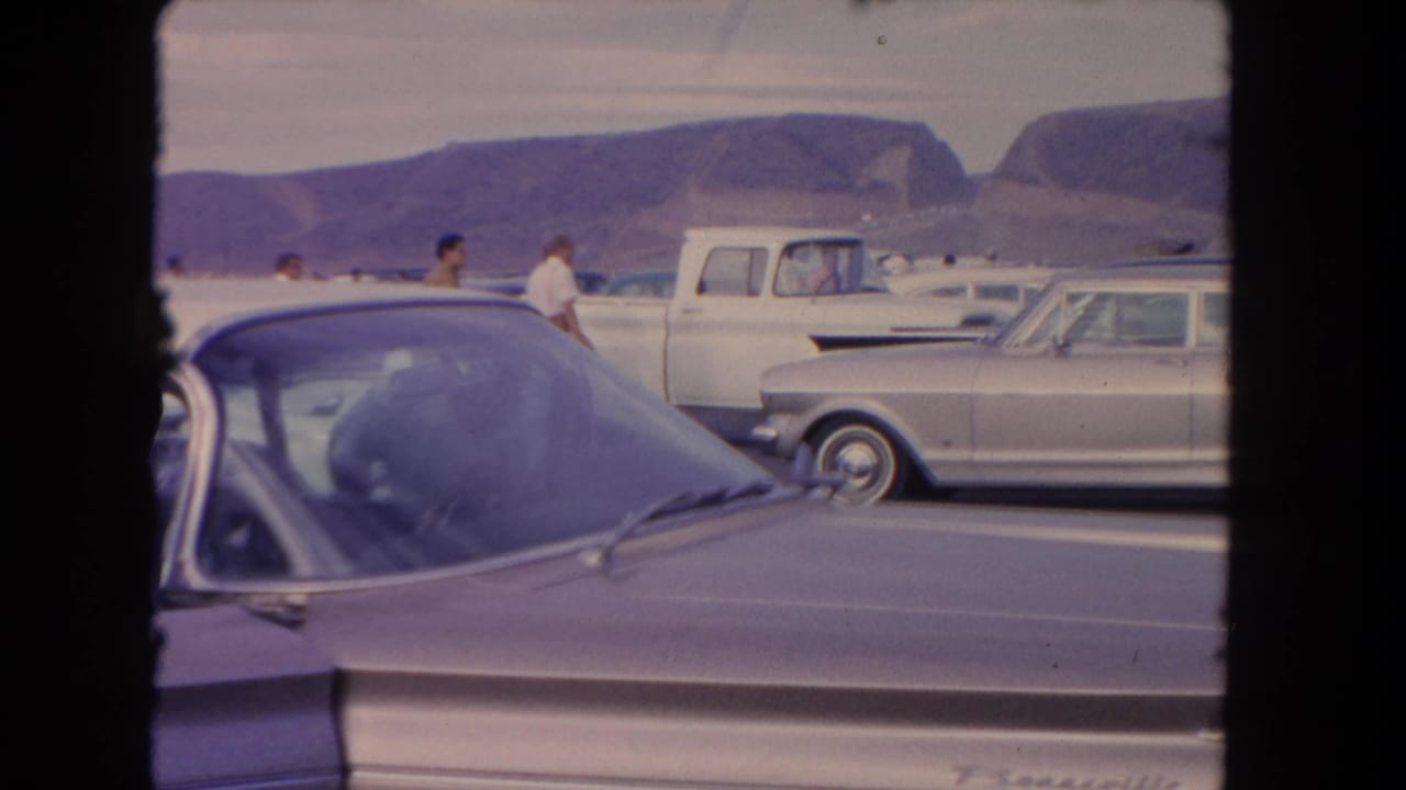 Foreground car and parked vehicles with people standing in a lot below distant mountains.