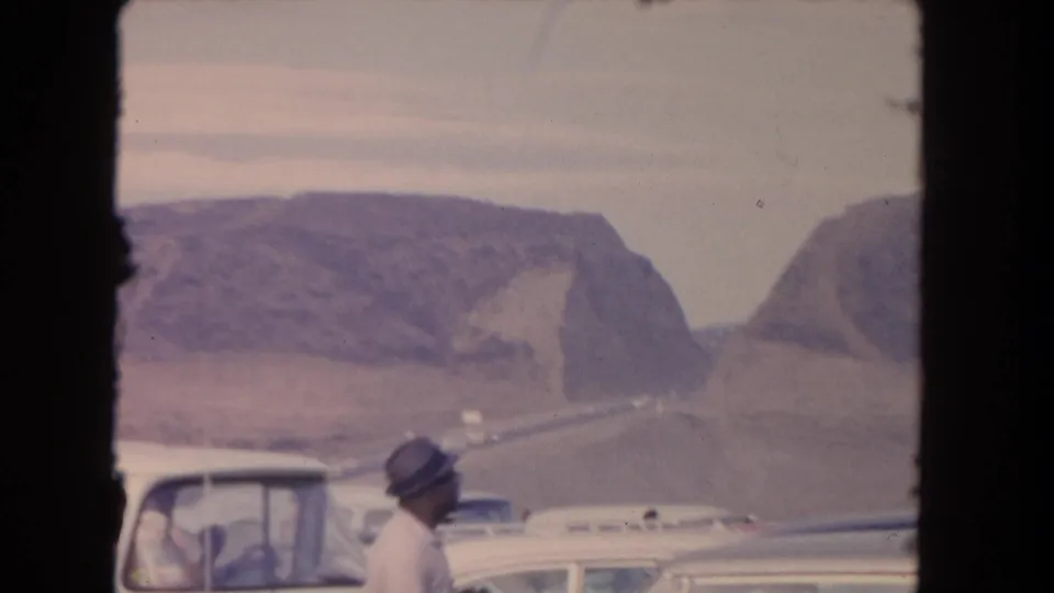 1962 Los Angeles-area frame with a man in a hat near parked classic cars, partial vehicle roofs, and mountains under a pale sky.